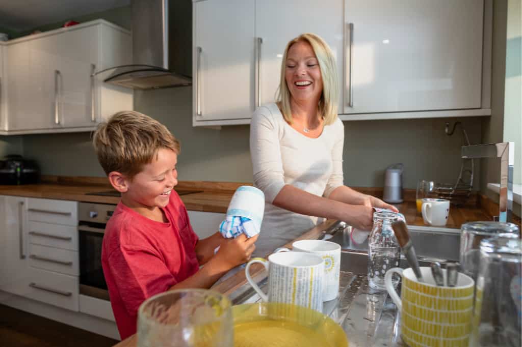 one of the ways to get kids to do chores is to have them help you with daily household activities like this boy helping wash dishes with his mom