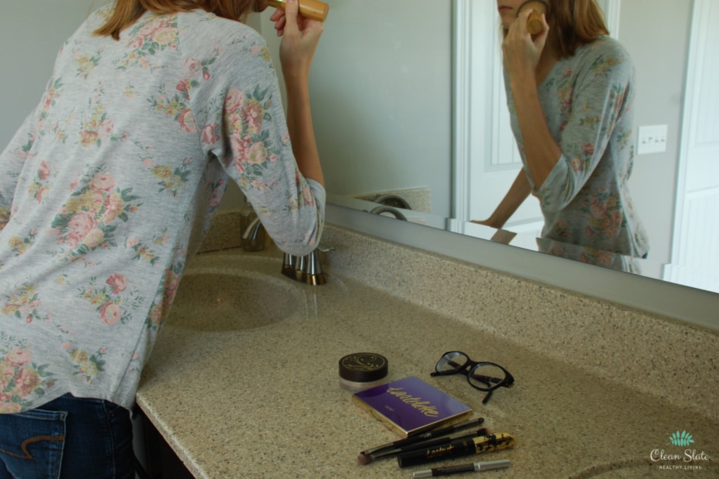 woman standing in front of bathroom mirror putting makeup on in the morning as part of her morning routine