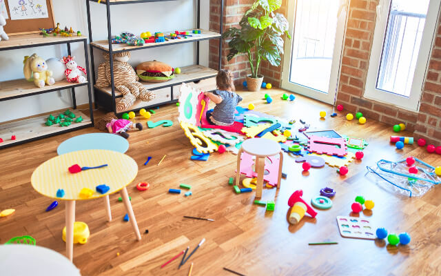 How to get kids to clean up. Girl is sitting in a messy room full of toys.