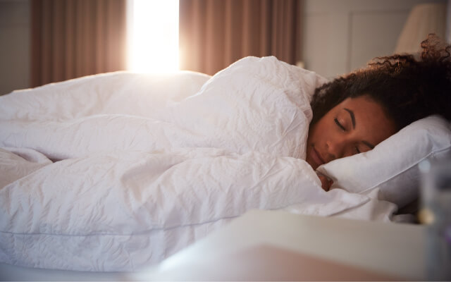 Woman is sleeping in her bed with sun shining in through the window. Getting good sleep is an important way to be more productive.