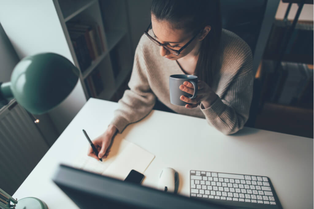 highly productive woman sitting at computer desk writing a list and drinking tea